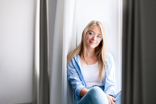 Young woman relaxing on windowsill at home. Happy girl enjoying fresh air and calmness. Concept of female mental health, slow living, social rest, mindfulness. Copy space