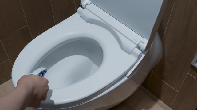 Hand of a person cleaning a white ceramic toilet bowl with a brush in a modern bathroom.