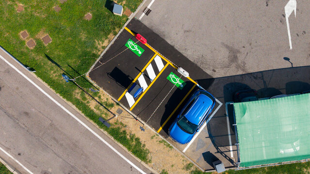 Aerial view of an electric vehicle charging station with a blue car parked in a marked bay, featuring green painted symbols and yellow lane markings in Milan, Lombardy, Italy.