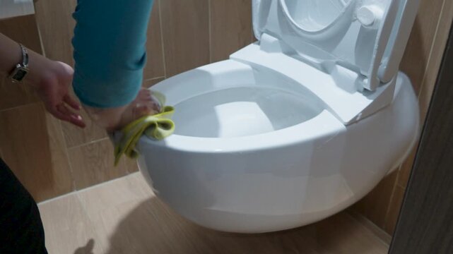 Woman cleaning and wiping a modern white ceramic toilet bowl with a cloth in the bathroom.