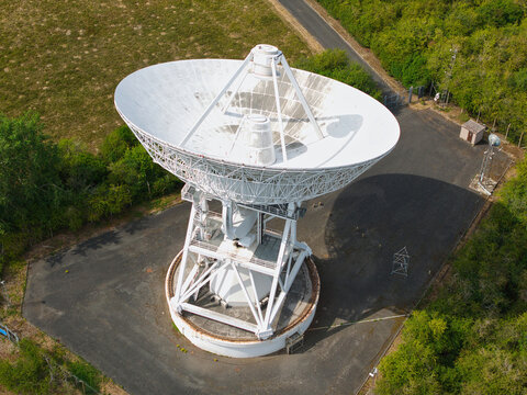Aerial view of the large white radio telescope dish at the Mullard Radio Astronomy Observatory, surrounded by green trees and paved ground in Harlton, England, United Kingdom.