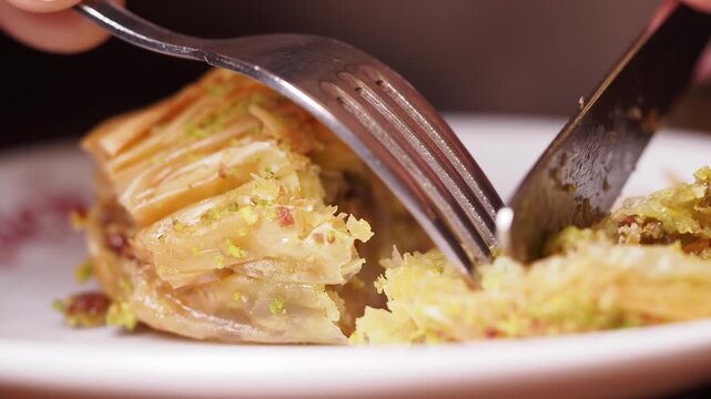Macro shot of cutting crispy baklava pastry with fork and knife on white plate