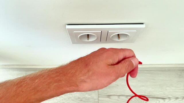 Close up view of male hands untangling a tangled red USB charging cable in front of a white double wall socket. Technology and power connection concept.