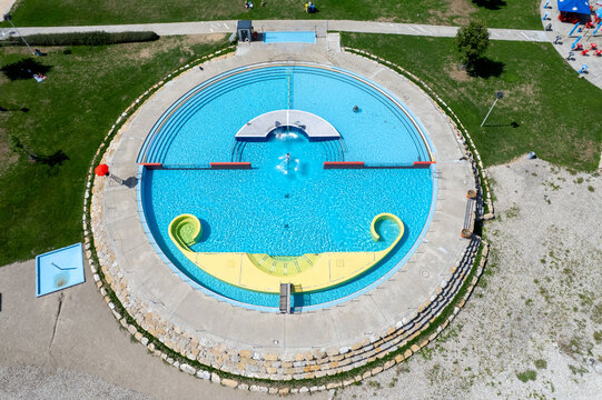 Aerial view of Piscines du Nid-du-Cro featuring a circular swimming pool with clear blue water, a distinctive yellow slide, and surrounding green lawns in Neuchatel, Neuchatel, Switzerland.