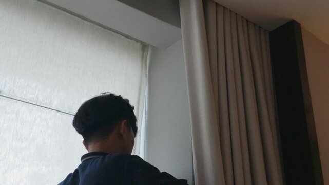 Back view of a young man standing near a window with curtains in a modern room.