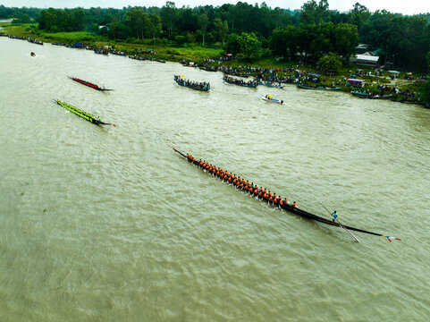 Aerial view of a traditional boat race with long wooden boats filled with rowers navigating the river near lush green banks in Manikganj, Dhaka Division, Bangladesh.