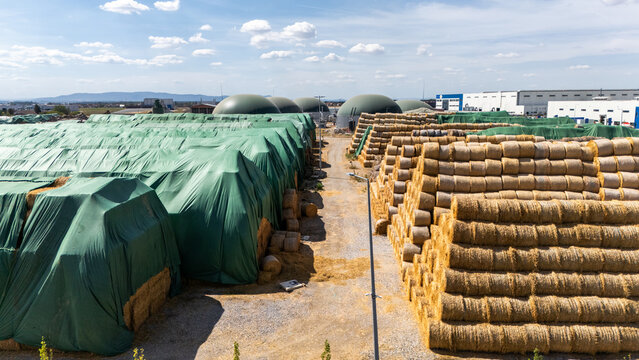 Aerial view of large stacks of hay bales, some covered with green tarps, alongside biogas digesters at an agricultural facility in Sabac, Vojvodina, Serbia.