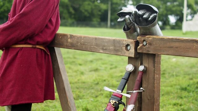 Medium shot captures medieval squire in red wool tunic standing near wooden fence where knight's steel plate gauntlets and sheathed swords rest before tournament combat...