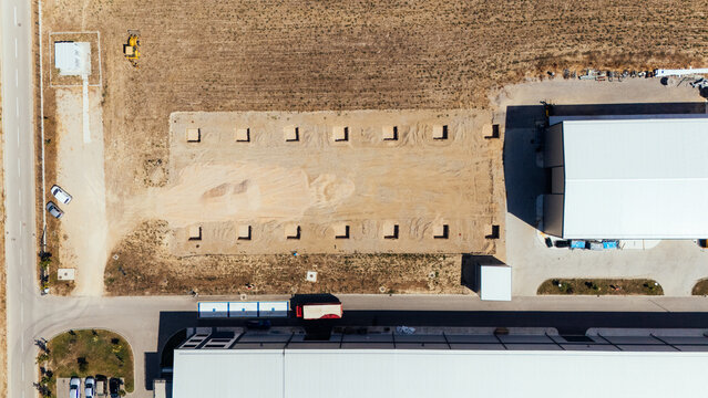 Aerial view of an industrial construction site with a large cleared dirt area, concrete foundation footings, and adjacent warehouse buildings in Sabac, Vojvodina, Serbia.
