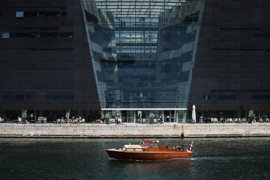Aerial view of a wooden boat cruising on the canal in front of the Black Diamond building in Copenhagen, Denmark.