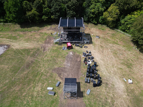 Aerial view of an outdoor concert stage setup in a grassy field with scattered equipment cases and surrounding trees in Valjevo, Kolubara District, Serbia.