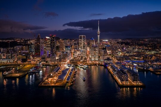 Illuminated Auckland city skyline and iconic Sky Tower glowing at night above the busy waterfront harbor in Auckland, New Zealand.