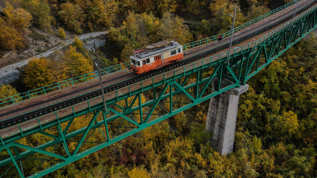 Aerial view of a train crossing a green metal railway bridge over a forest with autumn foliage in Montenegro.