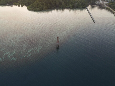 Aerial view of Waisai Harbour with a navigation beacon standing in the clear turquoise waters, surrounded by lush mangroves and a wooden pier in Raja Ampat, Papua Barat Daya, Indonesia.