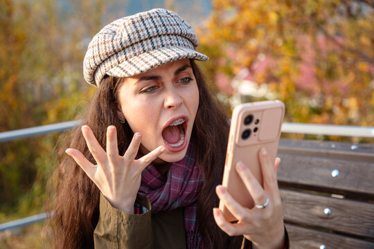 Young Caucasian woman holding smartphone and shouting in anger after reading unexpected online news. Aggression, frustration, and negative emotions concept.