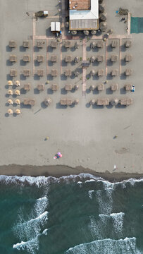 Aerial view of Ada Bojana beach with rows of thatched umbrellas, a restaurant building, and waves crashing on the sandy shore in Ulcinj Municipality, Montenegro.