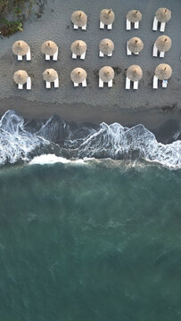 Aerial view of Ada Bojana beach featuring rows of straw umbrellas and sun loungers on dark sand with waves crashing along the shoreline in Ulcinj Municipality, Montenegro.