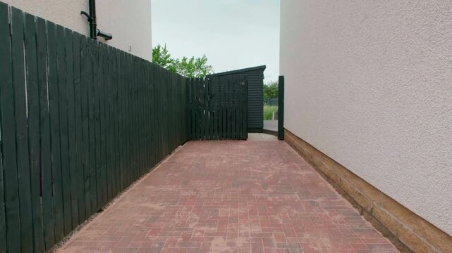 New Mono Driveway. Newly constructed red mono block brick driveway leads to a dark gate. Green fence lines the left side. White wall stands on the right. Driveway ends into a narrow entrance. Ideal fo