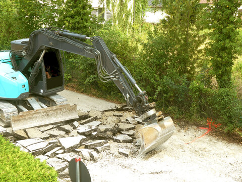 Aerial view of an excavator breaking up asphalt at a road construction site surrounded by trees in Garching, Bavaria, Germany.