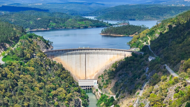 Aerial view of a large concrete arch dam spanning a river valley, surrounded by lush green forested hills and a vast reservoir in Portugal.