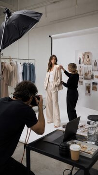 Studio fashion photoshoot with female model in beige tailored suit while stylist adjusts jacket and photographer captures images against white backdrop