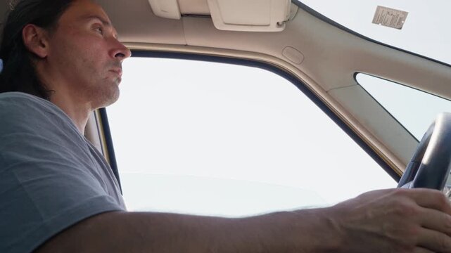 Driver gripping steering wheel inside car cabin, left-side profile with focused forward gaze and daylight rim lighting, with driver, man, hand, steering detail, showing wheel, grip, forearm, profile