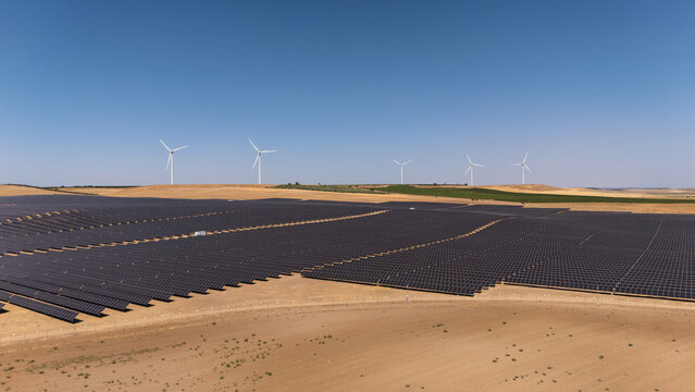 Aerial view of a large solar photovoltaic power plant with rows of solar panels and wind turbines in the background under a clear blue sky in Medina del Campo, Castile and Leon, Spain.