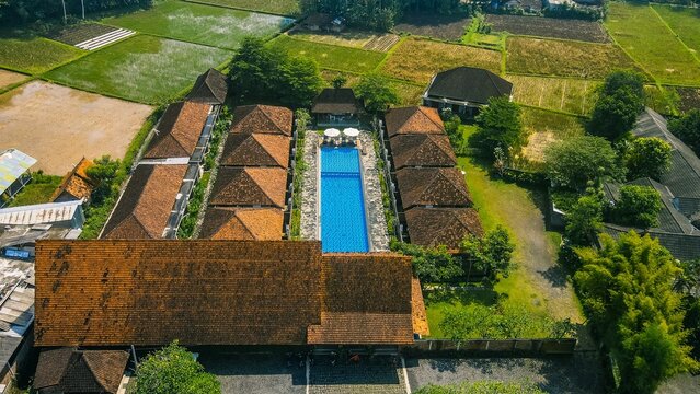 Aerial view of a resort complex with a long swimming pool surrounded by traditional terracotta-roofed villas and lush green rice fields in Borobudur, Central Java, Indonesia.