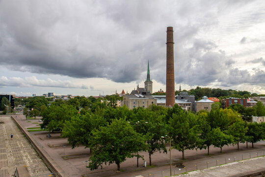 Tallinn cityscape view from the brutalist Linnahall roof under cloudy sky. Historic industrial brick chimney of Kultuurikatel and medieval St Olaf church spire rising over a green park.