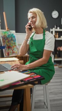 Woman painter in green apron talking on smartphone with hand on laptop amid easel and paints in studio; serene concentration.