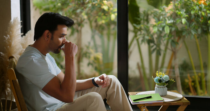 Indian calm young adult gen z relax man sitting on chair hold tea mug drink coffee enjoy day room male boy spend time take casual job break home house place