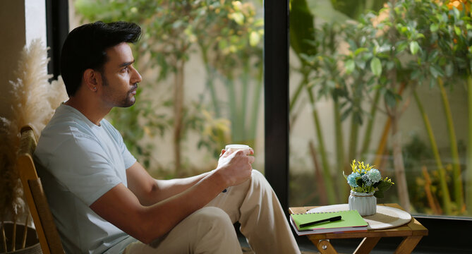 Indian calm young adult relax man sitting on chair hold tea mug coffee enjoy day room male boy spend time take casual job break home house place