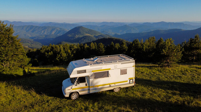 Aerial view of a white camper van parked on a grassy mountain slope surrounded by pine trees with distant mountain ranges in the background in Mokra Gora, Zlatibor District, Serbia.