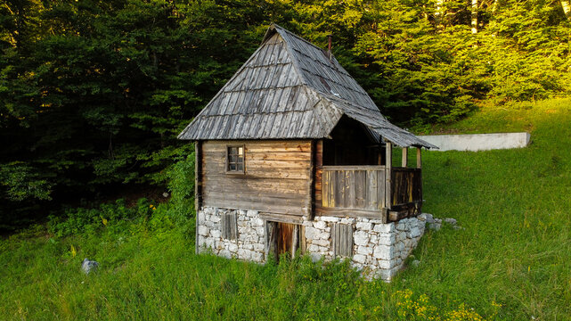 Aerial view of a traditional wooden cabin with a stone base nestled in a lush green meadow surrounded by dense forest in Mokra Gora, Zlatibor District, Serbia.