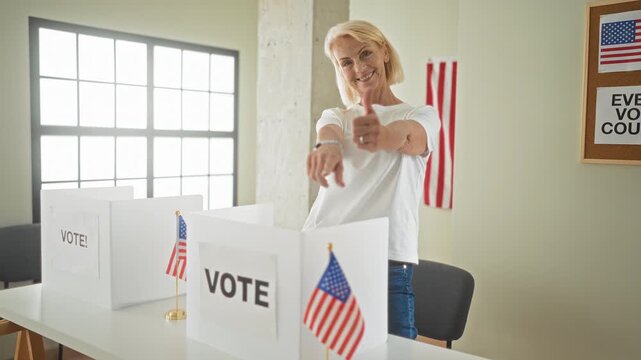 Middle age woman points finger at camera and gives thumbs up beside vote booth with small american flags in a building; civic pride.