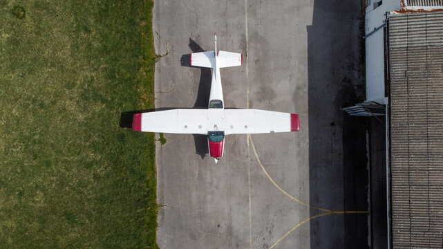 Aerial view of a small white and red light aircraft parked on the tarmac at Lisiciji Jarak Airport next to a hangar and grassy field in Padinska Skela, Grad Beograd, Serbia.
