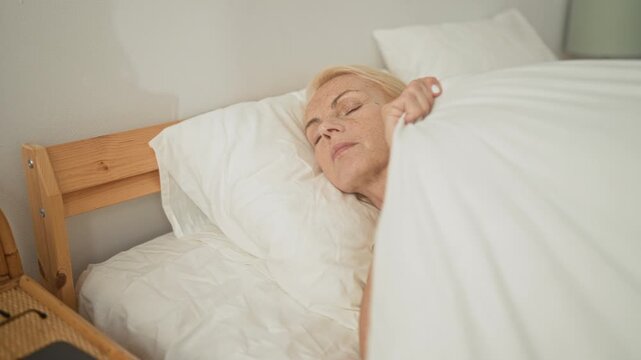 Woman middle age blonde sleeping while holding a white duvet, hand with painted nails visible on a wooden bed frame in a studio bedroom; peaceful.