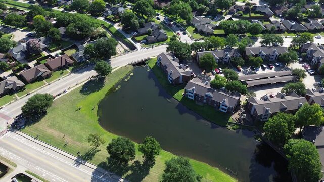 Reflective retention ponds flank Parkway Boulevard at Moore Road pedestrian crossings. Manicured grass slopes, brick apartment terraces, paved parking bays define scenic Coppell, Texas.