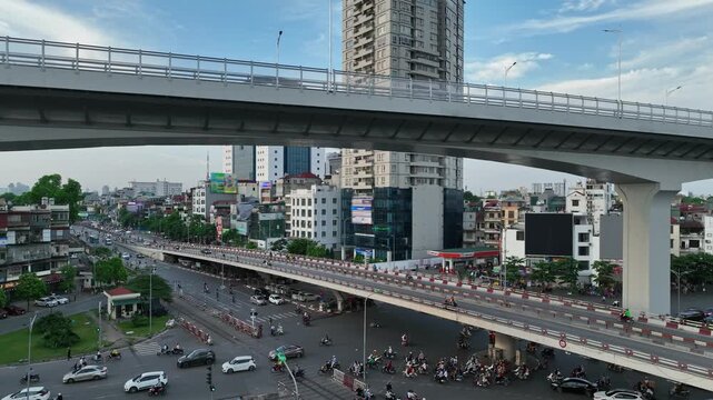 Urban traffic flows beneath Hanoi's elevated Ring Road 3 over Giai Phong Street, surrounded by dense buildings, motorcycles, cars, buses, and city infrastructure.