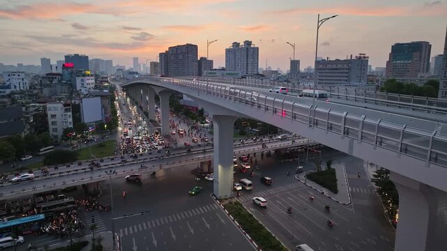 Urban traffic flows beneath Hanoi's elevated Ring Road 3 over Giai Phong Street at sunset, surrounded by apartment buildings and city lights.