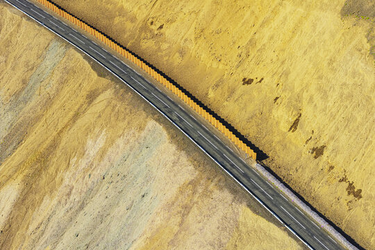 Aerial view of a paved road cutting through a barren, golden-hued volcanic landscape with a tall orange barrier fence in Oceanside, Mulathing, Iceland.