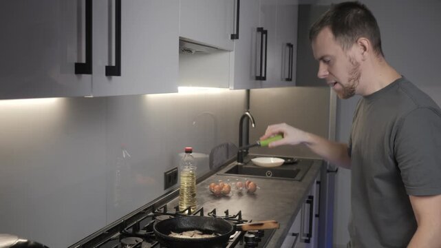 Man handling eggs beside gas cooktop in modern kitchen with cast iron skillet, open carton, oil bottle, white bowl, reflective backsplash, and focused preparation for cooking