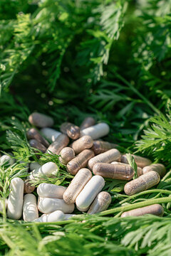 Close-up of white and clear herbal capsules with dietary supplements scattered, fresh green carrot tops. Healthy lifestyle, vegan vitamins, naturopathy, sports nutrition, and organic medical products