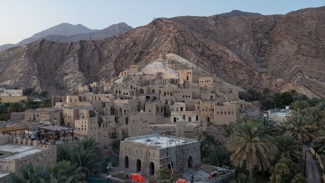 Aerial view of the historic mud-brick ruins of Birkat Al Mouz nestled against a rugged mountain backdrop, surrounded by lush date palm groves in Birkat Al Mouz, Ad Dakhiliyah Govern, Oman.