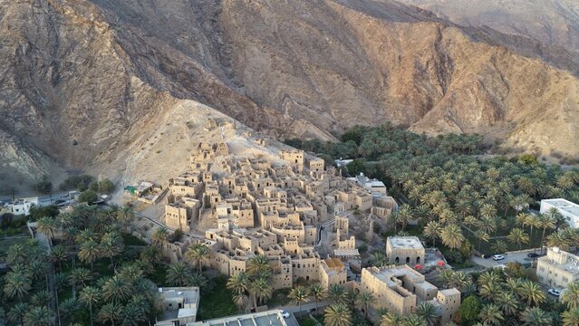 Aerial view of the ancient mud-brick ruins of Birkat Al Mouz surrounded by a lush date palm oasis and rugged mountain landscape in Birkat Al Mouz, Ad Dakhiliyah, Oman.