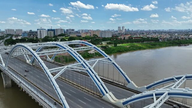Aerial view of Dong Tru Bridge crossing the Red River, with distinctive blue arches, busy roadway, riverside greenery, and Hanoi city skyline under a sunny sky.