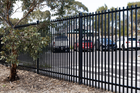 A black steel palisade security fence bordering a suburban car park in Australia, with parked vehicles visible through the fence rails. Commercial boundary or institutional perimeter fencing