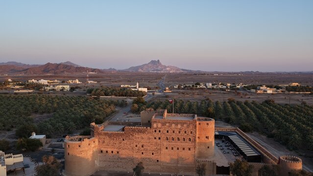 Aerial view of Jabreen Castle, a historic fortress with stone walls and towers, surrounded by date palm plantations and arid desert landscape in Bahla, Ad Dakhiliyah, Oman.