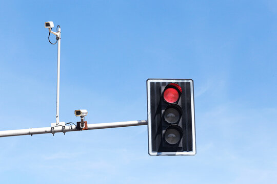 Red traffic light glows beside several road surveillance cameras against a clear blue sky. Urban traffic control background with stop signal monitoring equipment road safety and intersection.