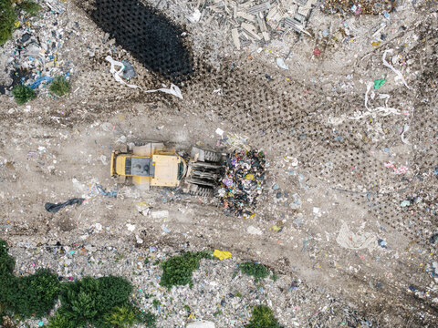 Aerial view of a yellow bulldozer moving a pile of waste at a landfill site with visible tire tracks and surrounding greenery in Laidzes pagasts, Talsu novads, Latvia.